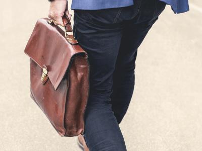 Eine Person in dunkler Hose und blauem Sakko geht auf einem hellen Untergrund und trägt eine braune Ledertasche in der linken Hand. Die Aufnahme zeigt den unteren Rücken- und Beinbereich sowie die Tasche aus der Nähe.