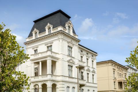 Weiße historische Stadtvilla im Gründerzeitstil mit aufwendig verzierter Fassade, Säulenbalkon und Mansarddach unter blauem Himmel, umgeben von grünen Bäumen.