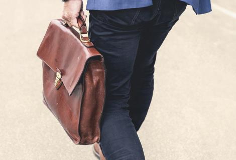 Eine Person in dunkler Hose und blauem Sakko geht auf einem hellen Untergrund und trägt eine braune Ledertasche in der linken Hand. Die Aufnahme zeigt den unteren Rücken- und Beinbereich sowie die Tasche aus der Nähe.