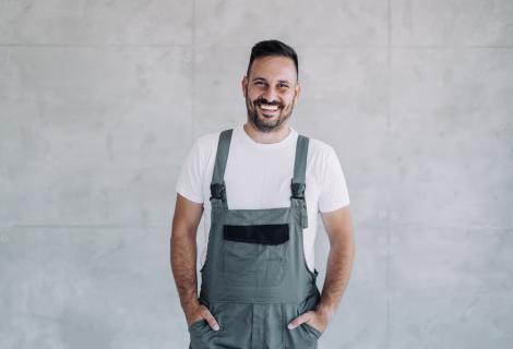 Ein lächelnder Mann in grauer Latzhose und weißem T-Shirt steht vor einer hellgrauen Betonwand und hat die Hände in den Hosentaschen.